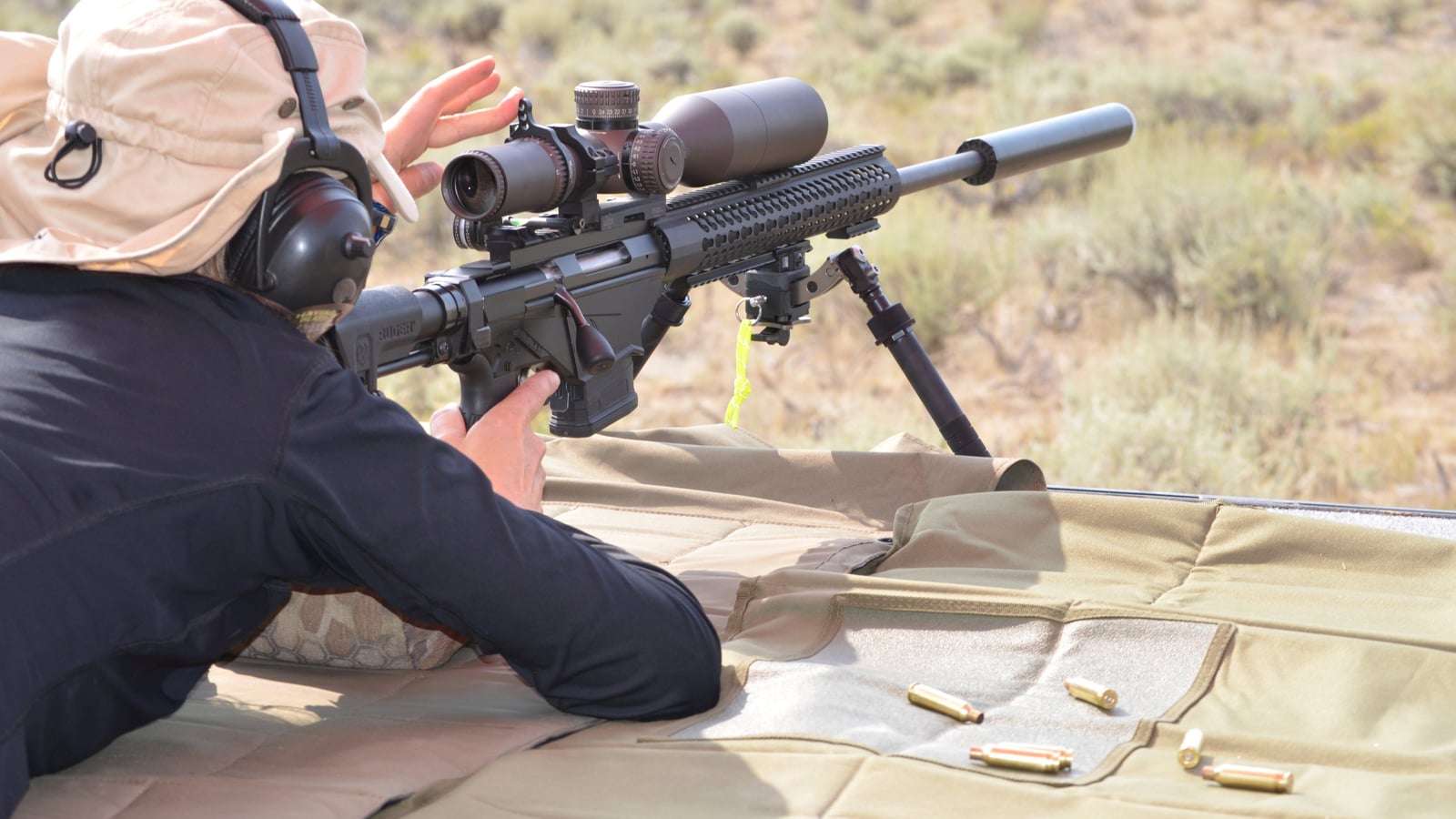 Shooter in prone position with a scoped rifle mounted on a bipod, at an outdoor shooting range