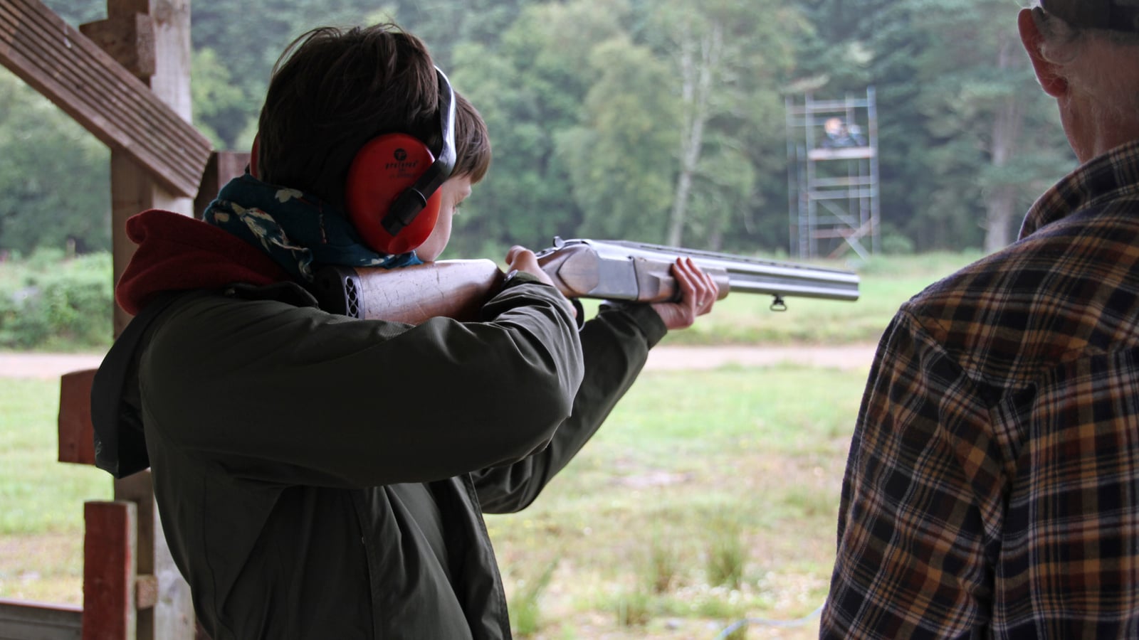 Shooter mounting a shotgun on an outdoor clay target firing line, under an instructor's supervision