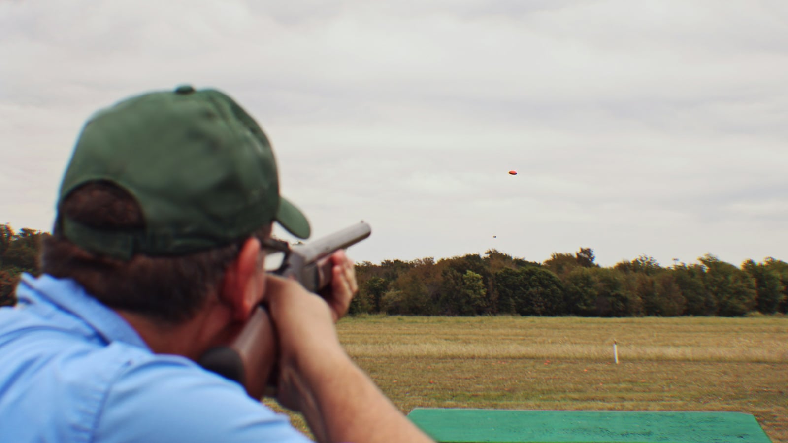 Shooter shouldering a shotgun at an outdoor firing point, with two clay targets visible in simultaneous flight in the sky above the treeline