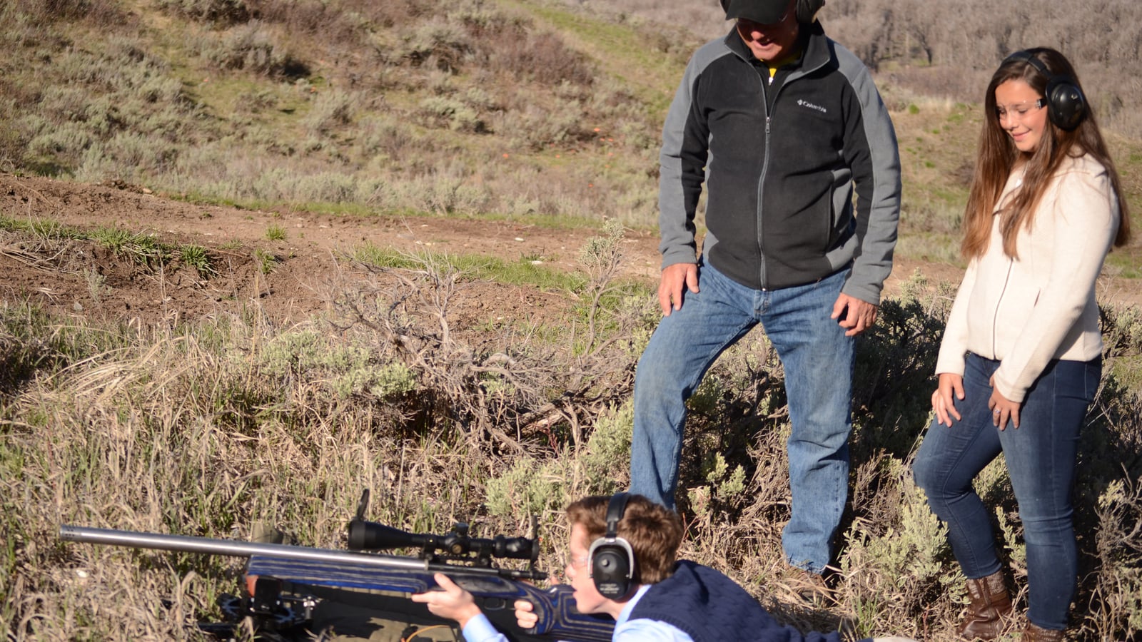 Shooter lying prone aiming at long range with a rifle on a bipod, family nearby outdoors