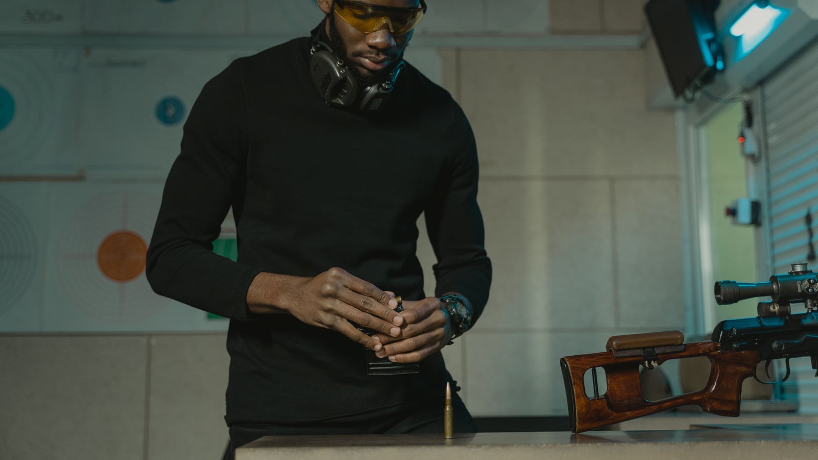 Shooter in dark clothing, safety glasses and hearing protection around the neck, loading a magazine at an indoor firing line in front of a rifle resting on the counter