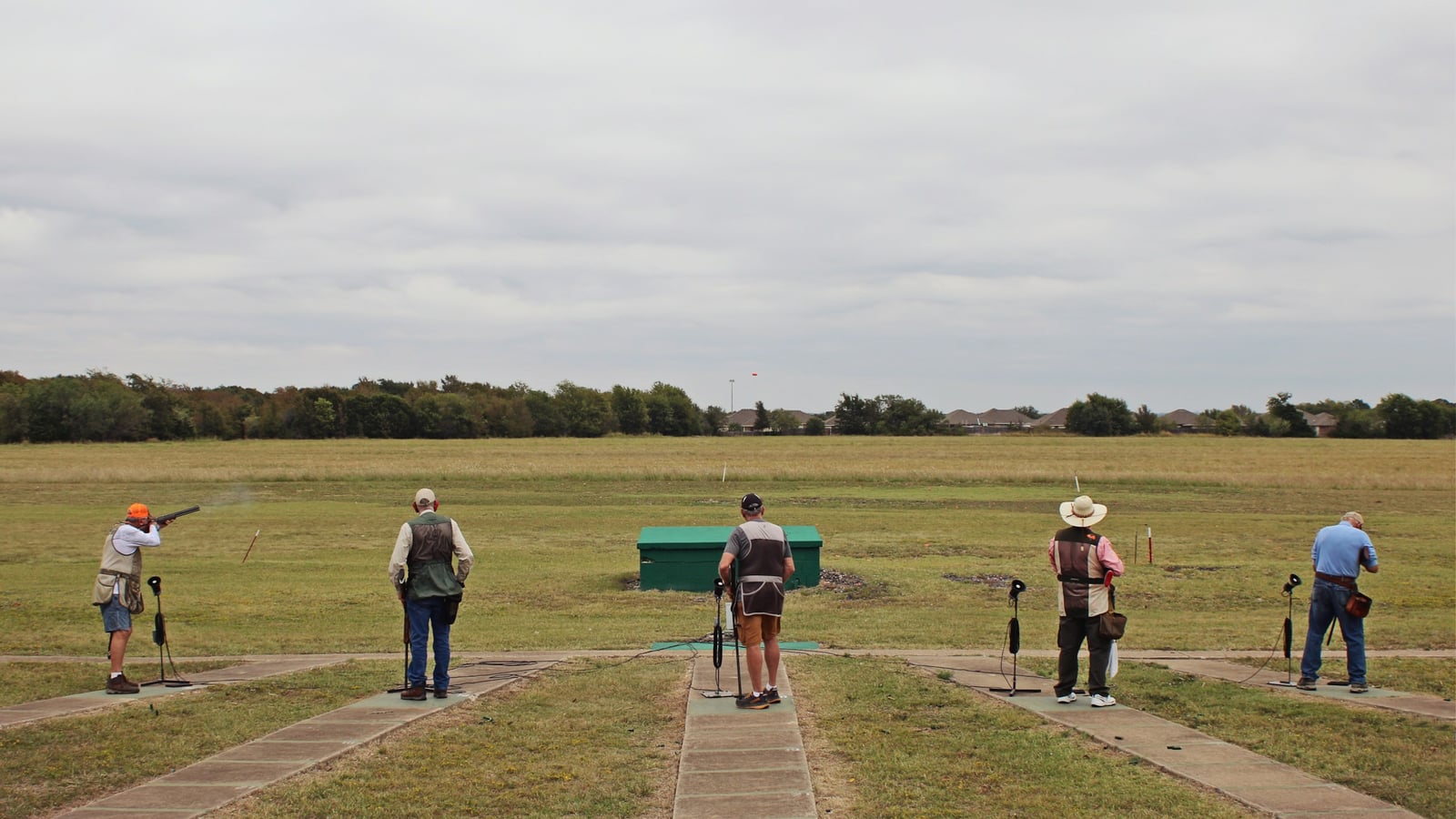 Five shooters lined up at their stations during a clay target event at a club, with the launch house at the center of the shooting line