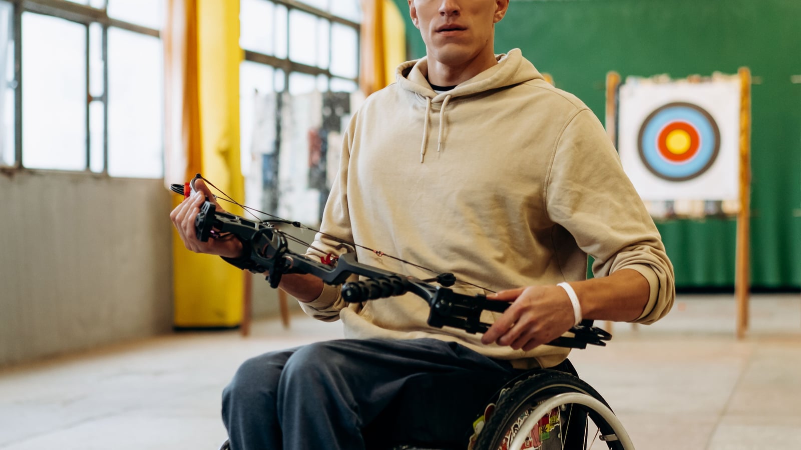 Athlete in a wheelchair holding a compound bow with an adapted grip, facing an official target in a covered shooting range
