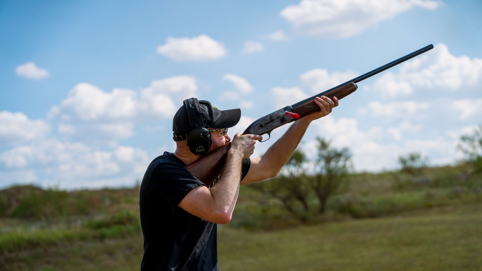 Tireur en protections auditives et oculaires épaulant un fusil basculant vers le ciel, dans un champ herbeux à ciel bleu