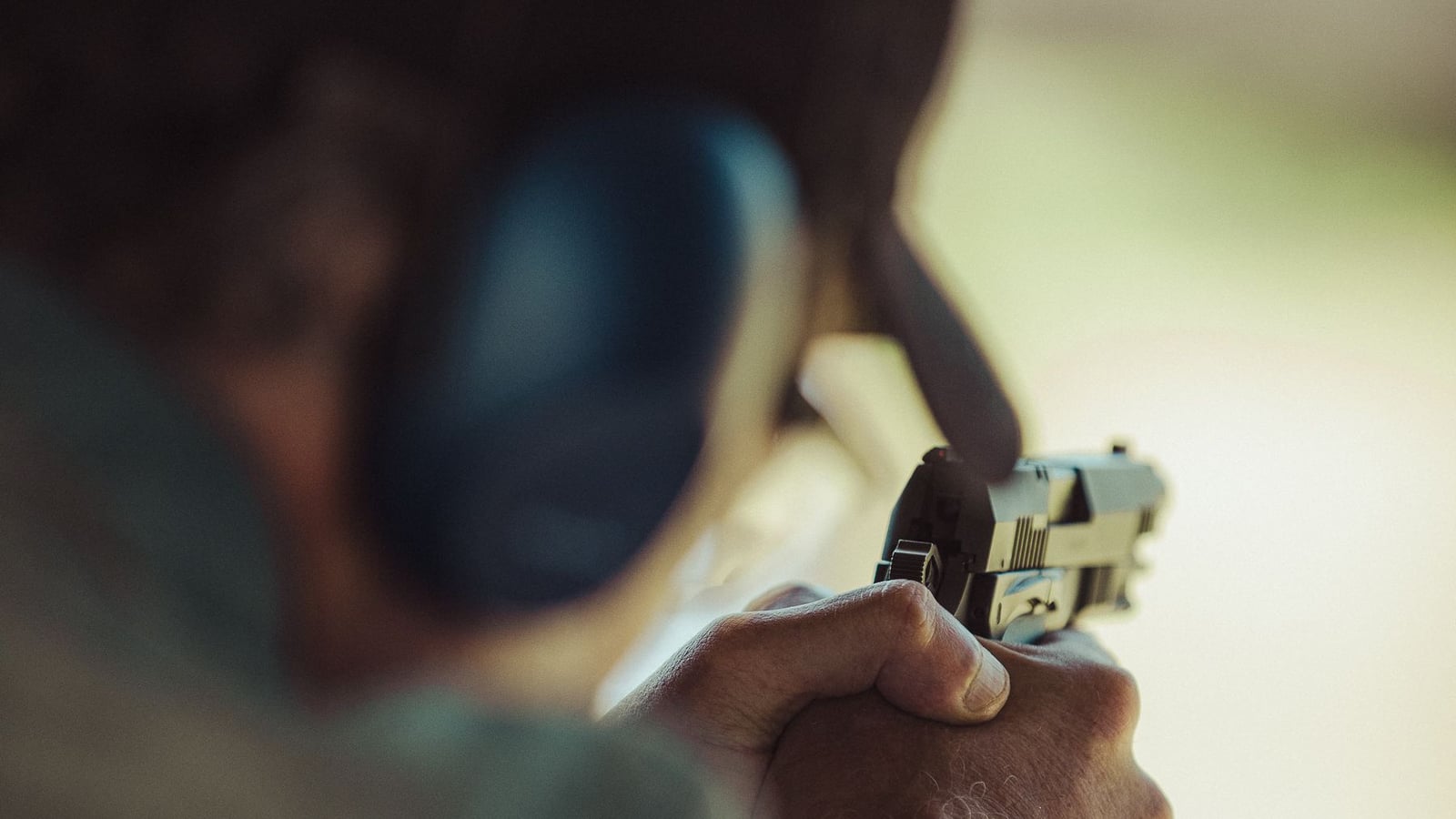 Shooter with extended arm holding a pistol at an outdoor range, wearing ear protection