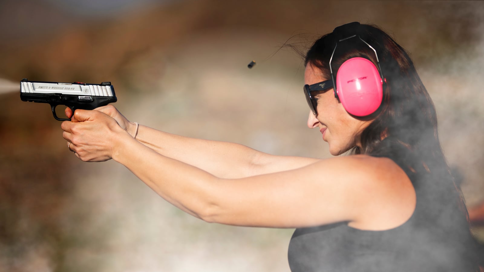 Female shooter in profile wearing glasses and pink hearing protection, at the moment of firing a pistol outdoors