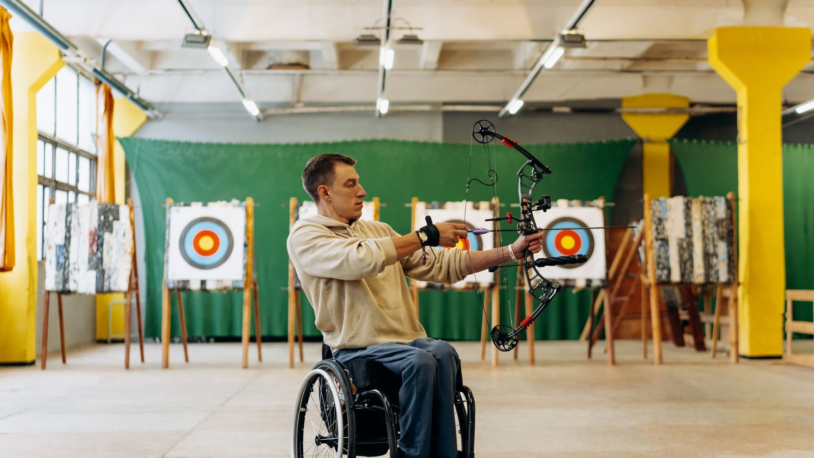Athlete in a wheelchair drawing a compound bow facing targets in a bright, colorful archery club