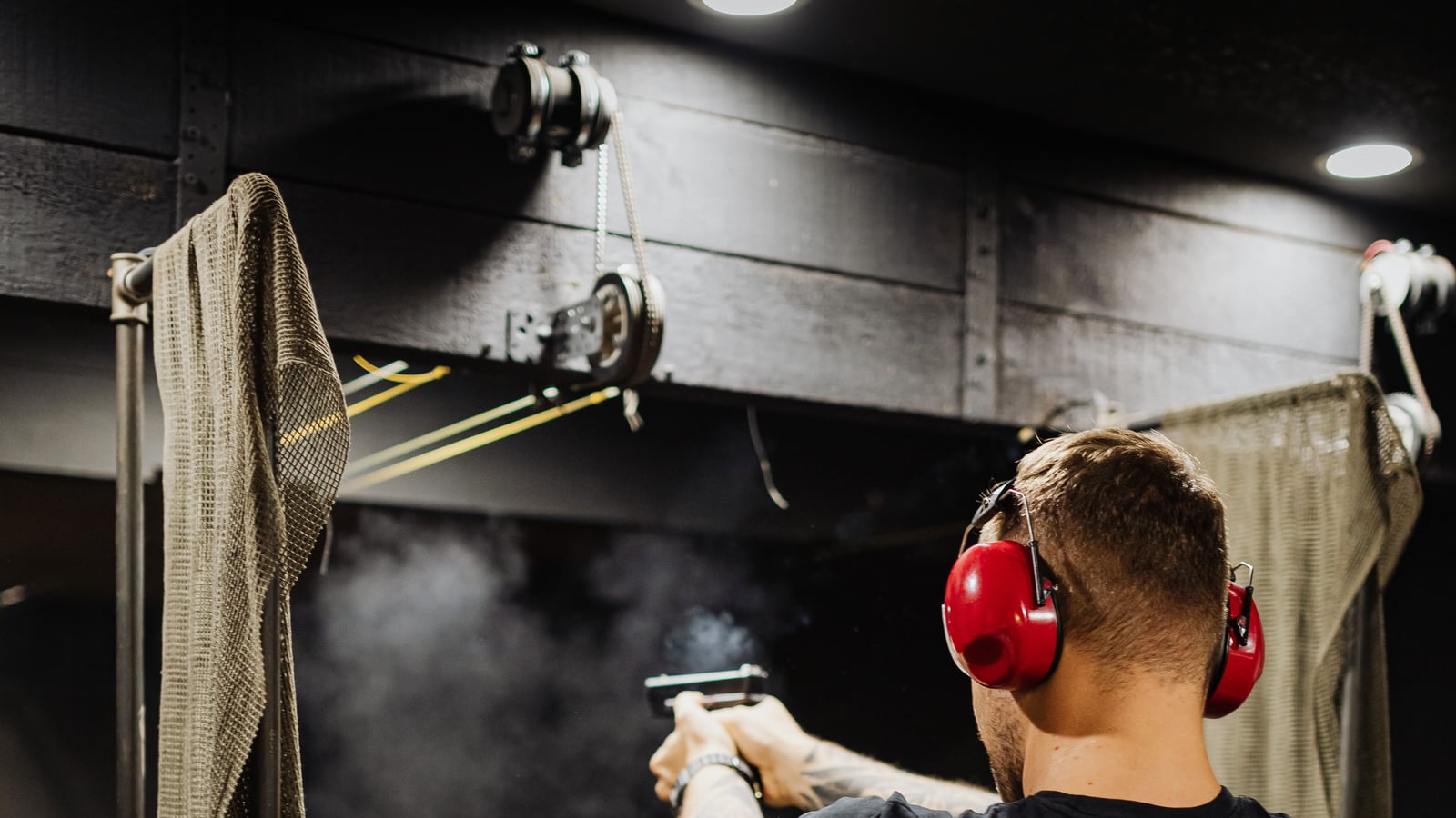 Shooter seen from behind, red hearing protection, firing a pistol on a covered range